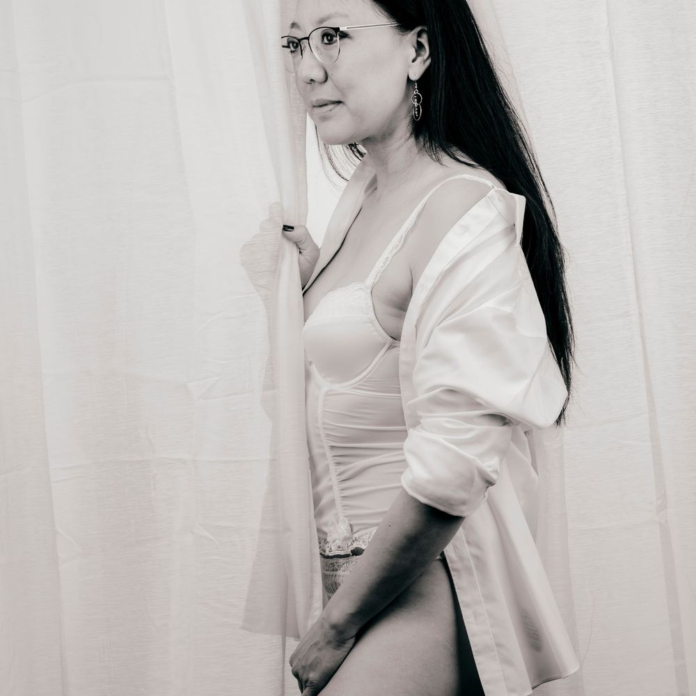 Woman in white lingerie and open shirt poses elegantly beside sheer curtains in black and white photo.