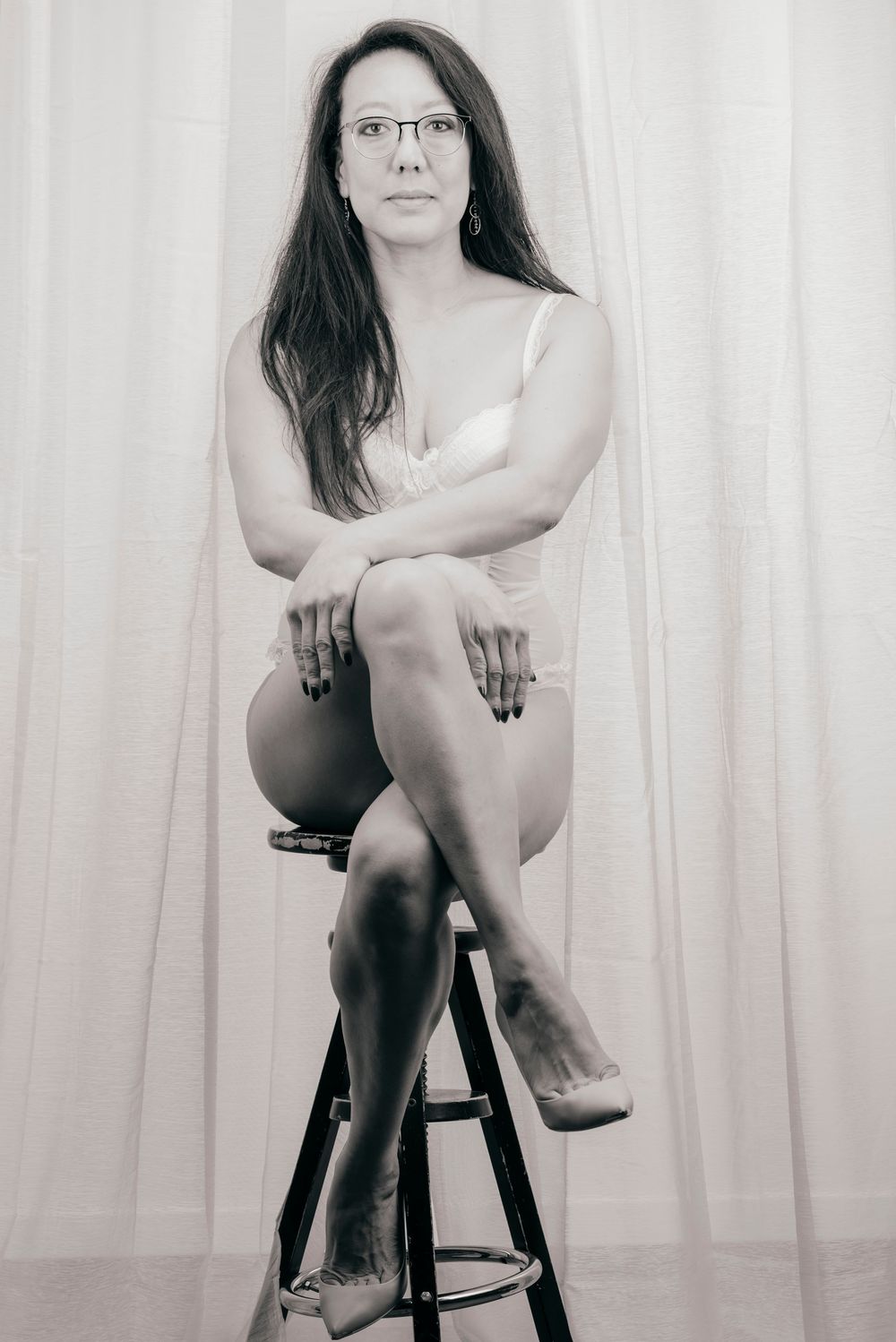 Woman in white lingerie sits gracefully on stool before white curtain in classic black and white portrait.