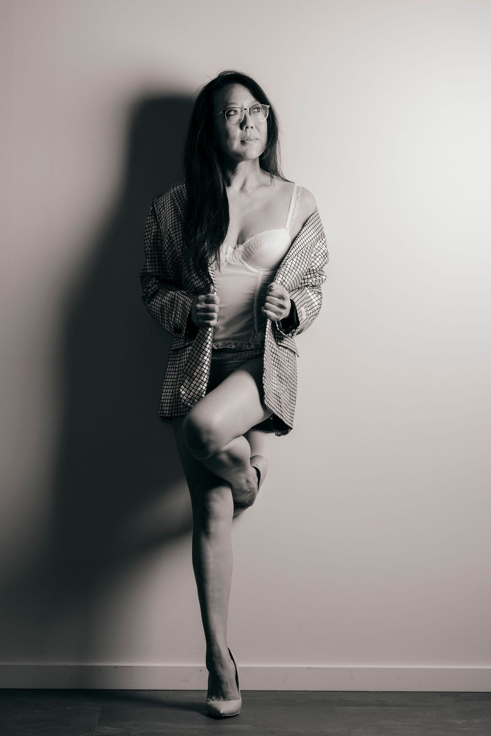 Woman in lingerie and oversized blazer poses confidently against wall in black and white boudoir photo.
