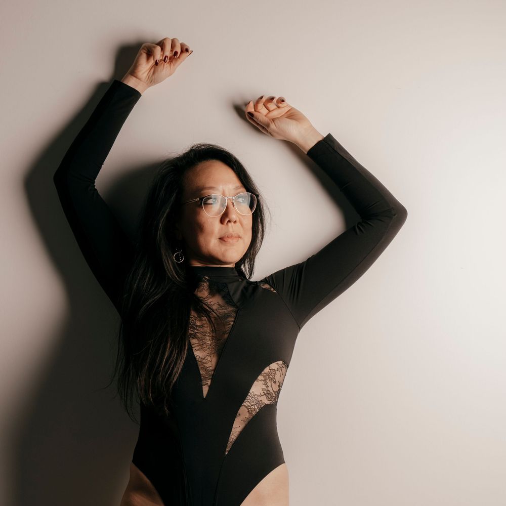Woman in black lace bodysuit poses dynamically with arms raised against light gray studio background.