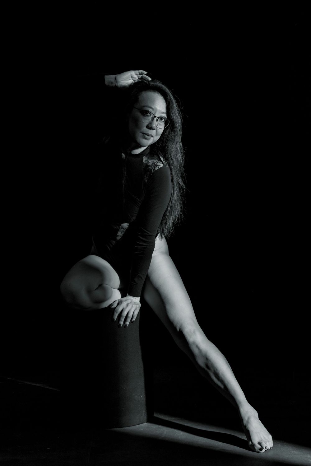 Dramatic black and white boudoir photo of woman in black bodysuit posing on pedestal in dark studio.