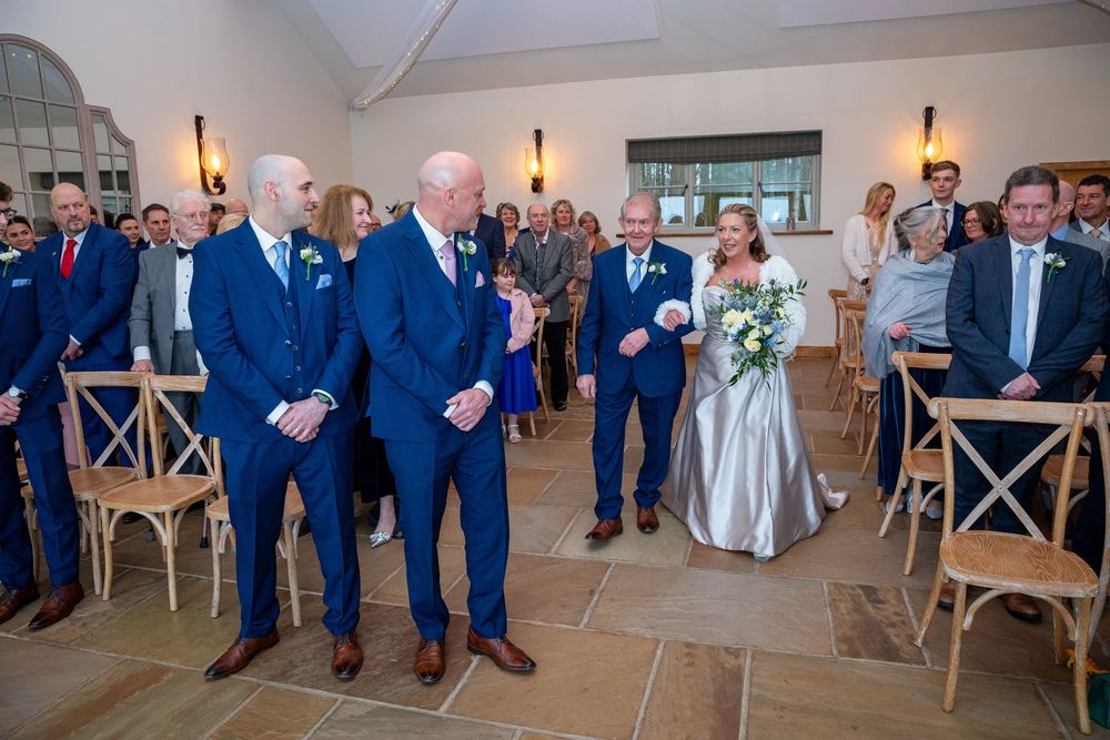 Bride walks down the aisle with escort at elegant wedding ceremony while guests stand in admiration.