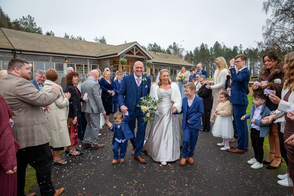 Newlywed couple walks through confetti shower surrounded by cheering wedding guests outdoors.