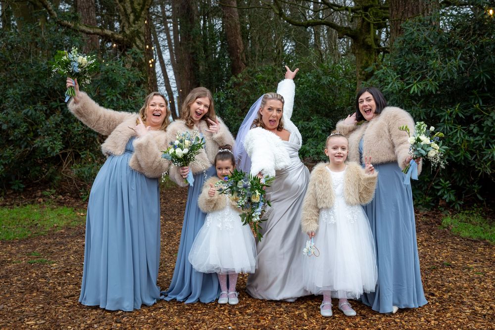 Bride poses joyfully with bridesmaids in blue dresses and flower girls in autumn woodland setting.