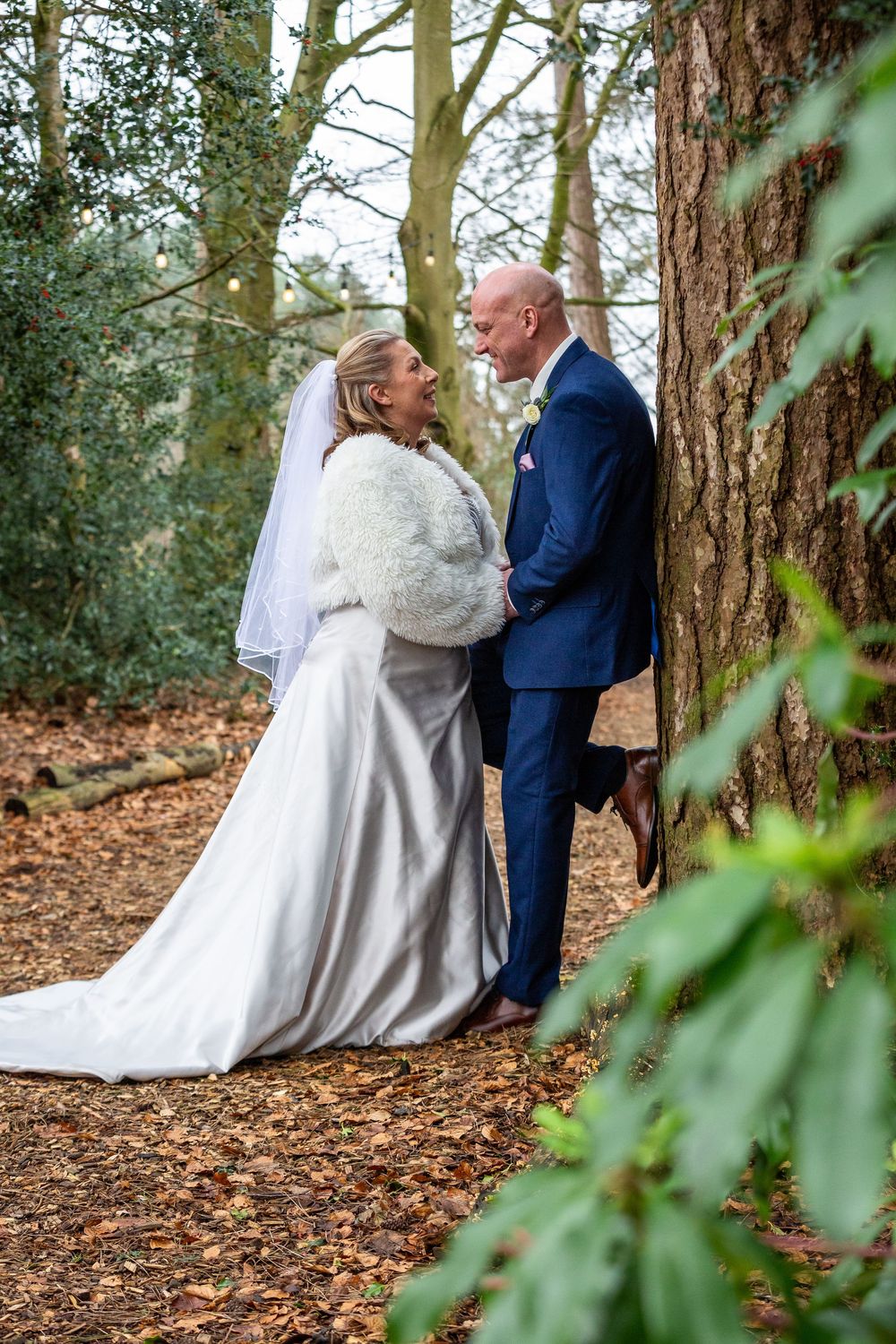 Bride and groom share romantic moment leaning against tree in woodland wedding photo.
