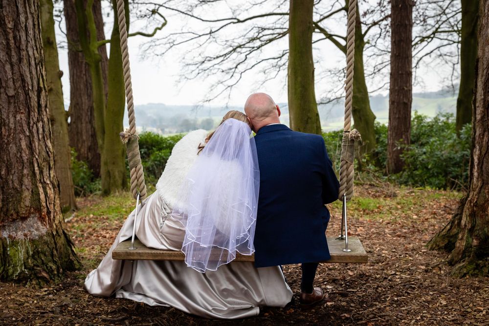 Bride and groom sit together on wooden swing surrounded by tall trees at wedding venue.