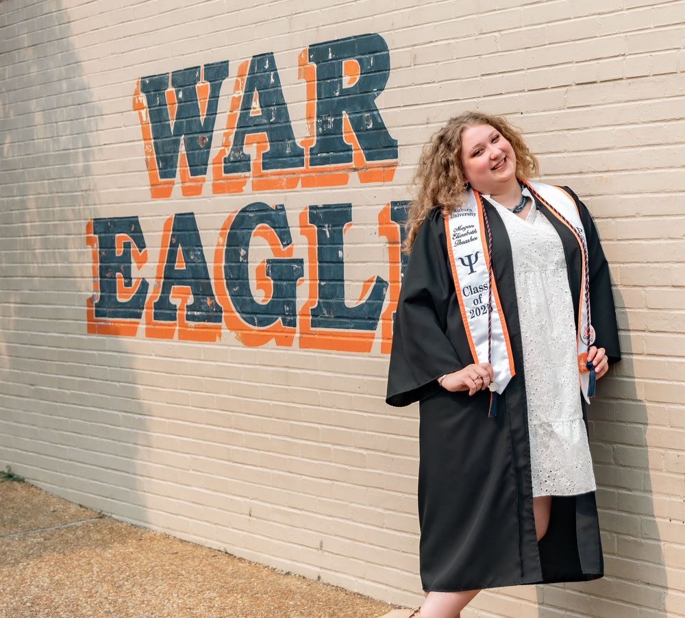 Graduate in cap and gown poses against brick wall with orange War Eagle text painted on it.