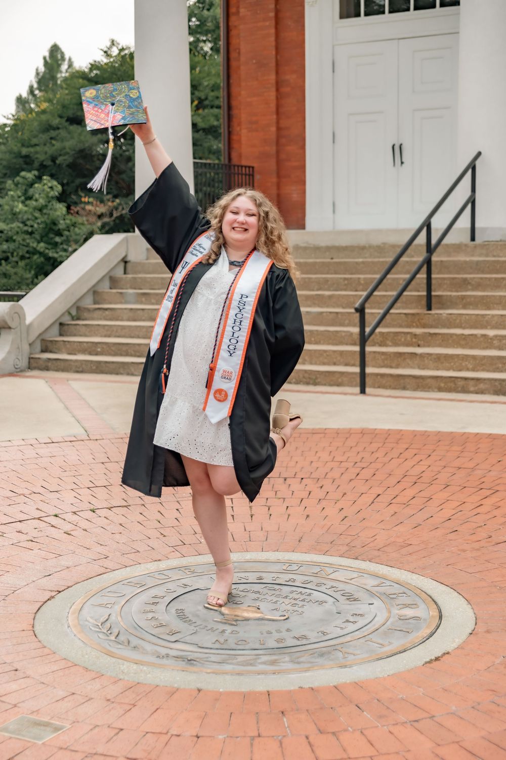 Graduate poses on commemorative university seal set in brick courtyard wearing cap and gown against brick building.