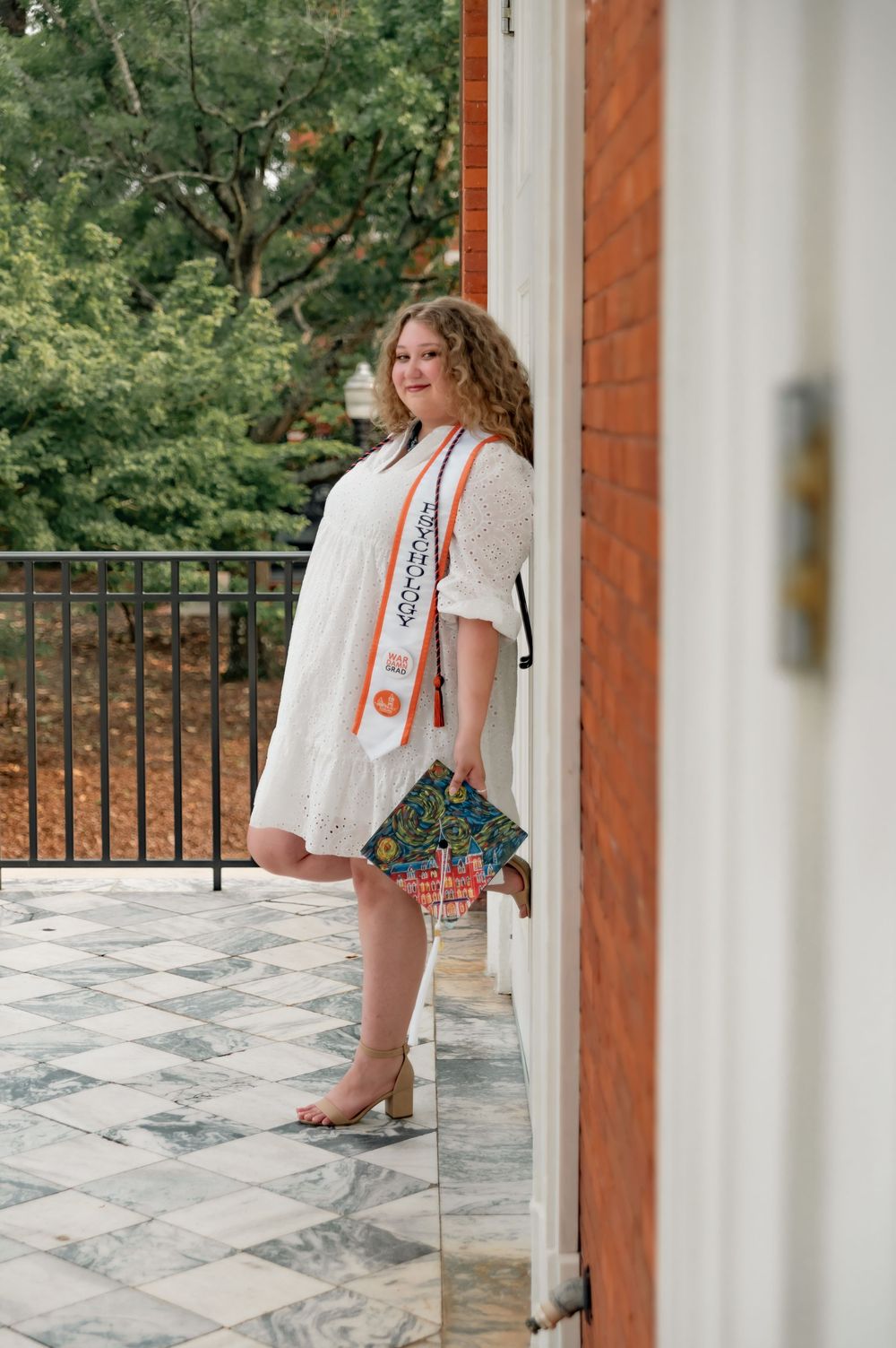 A woman poses on a porch in a white embroidered dress with a patterned handbag near brick pillars and greenery.