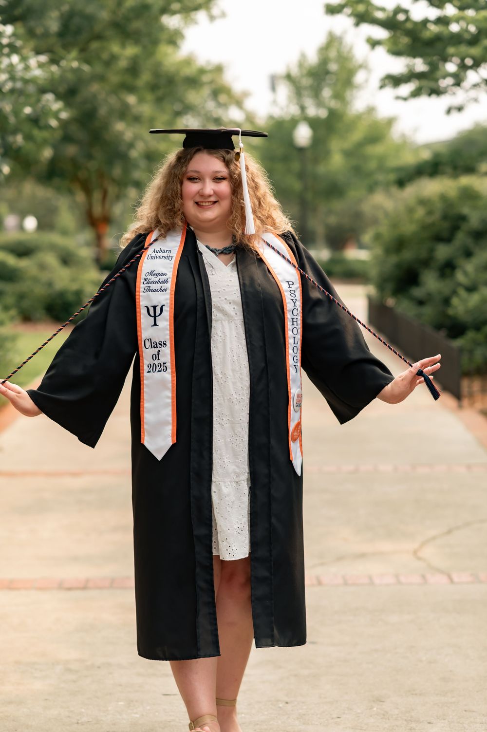 Graduate in black cap and gown with decorative sash poses outdoors on pathway during commencement ceremony.