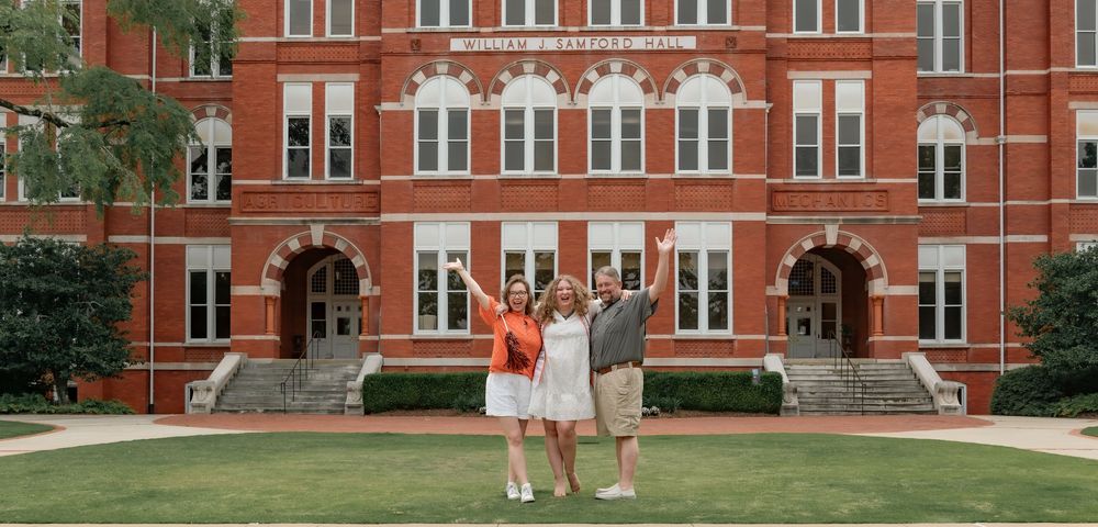 Classic red brick college dormitory building with white trim and arched entryways stands among green trees on campus.