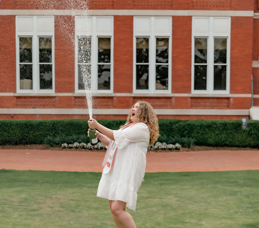A person in a white dress stands on a lawn holding a graduation cap in front of a brick building with large windows.