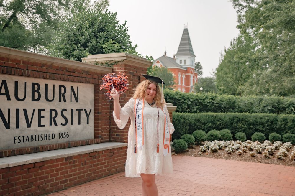 Graduate in white dress celebrates at Auburn University's iconic brick entrance sign with Samford Hall tower in background.