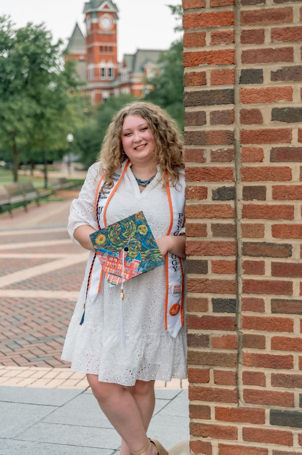 A person in a white eyelet dress and orange lanyard stands next to a brick wall on a college campus walkway.