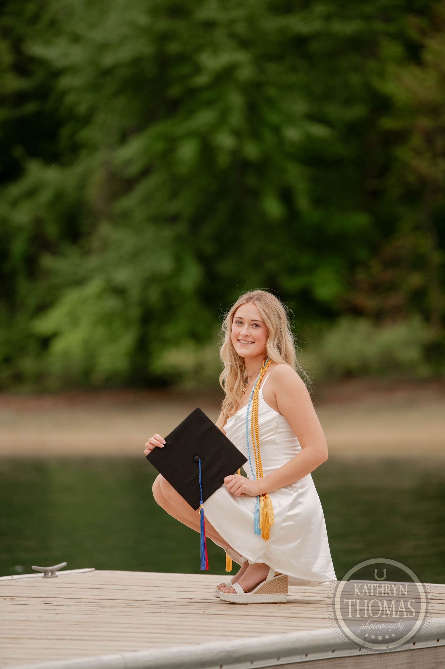 Graduate in white dress poses with diploma and stole on a wooden dock by a serene lake during sunset.