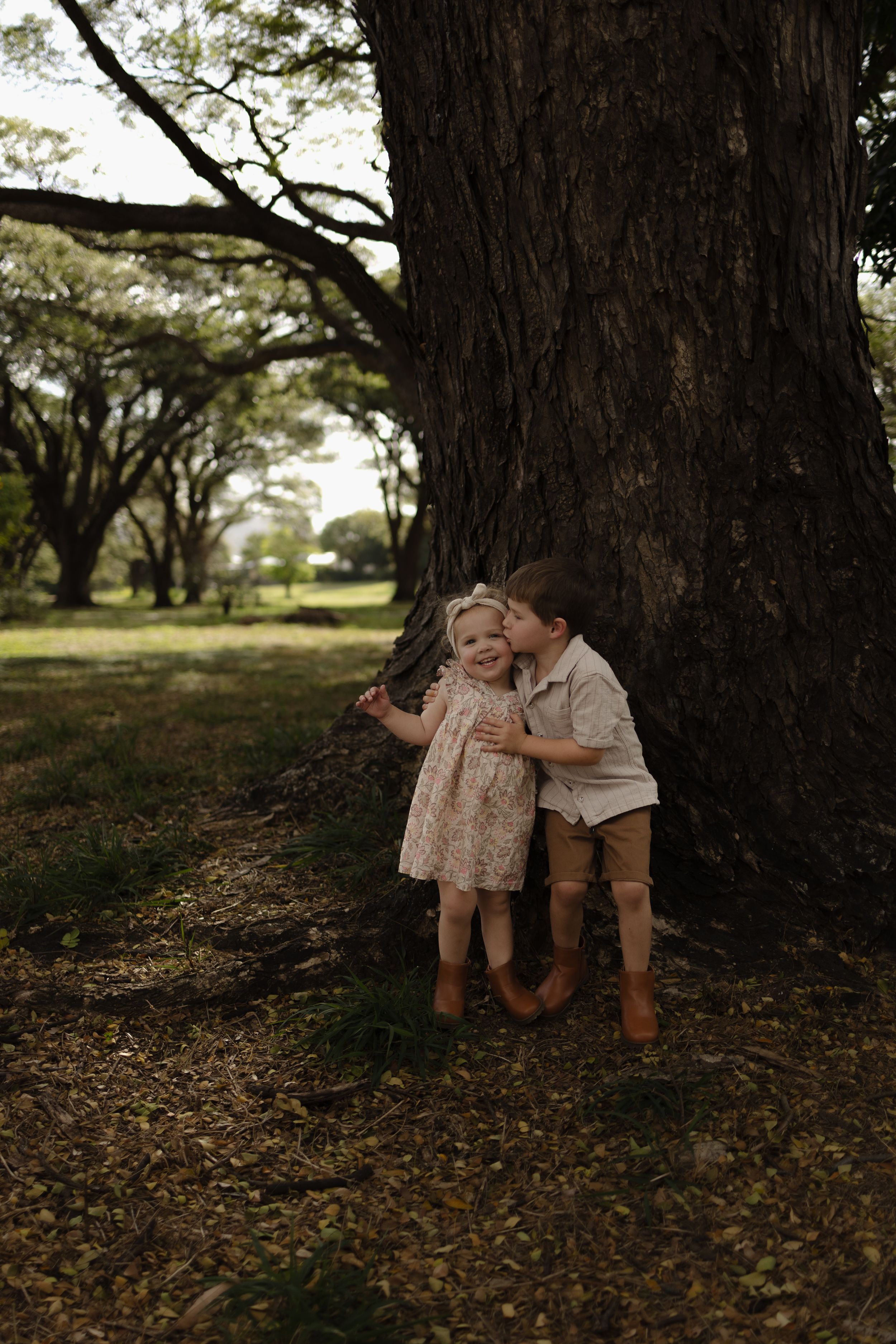 Two young children share a sweet moment under a large tree.