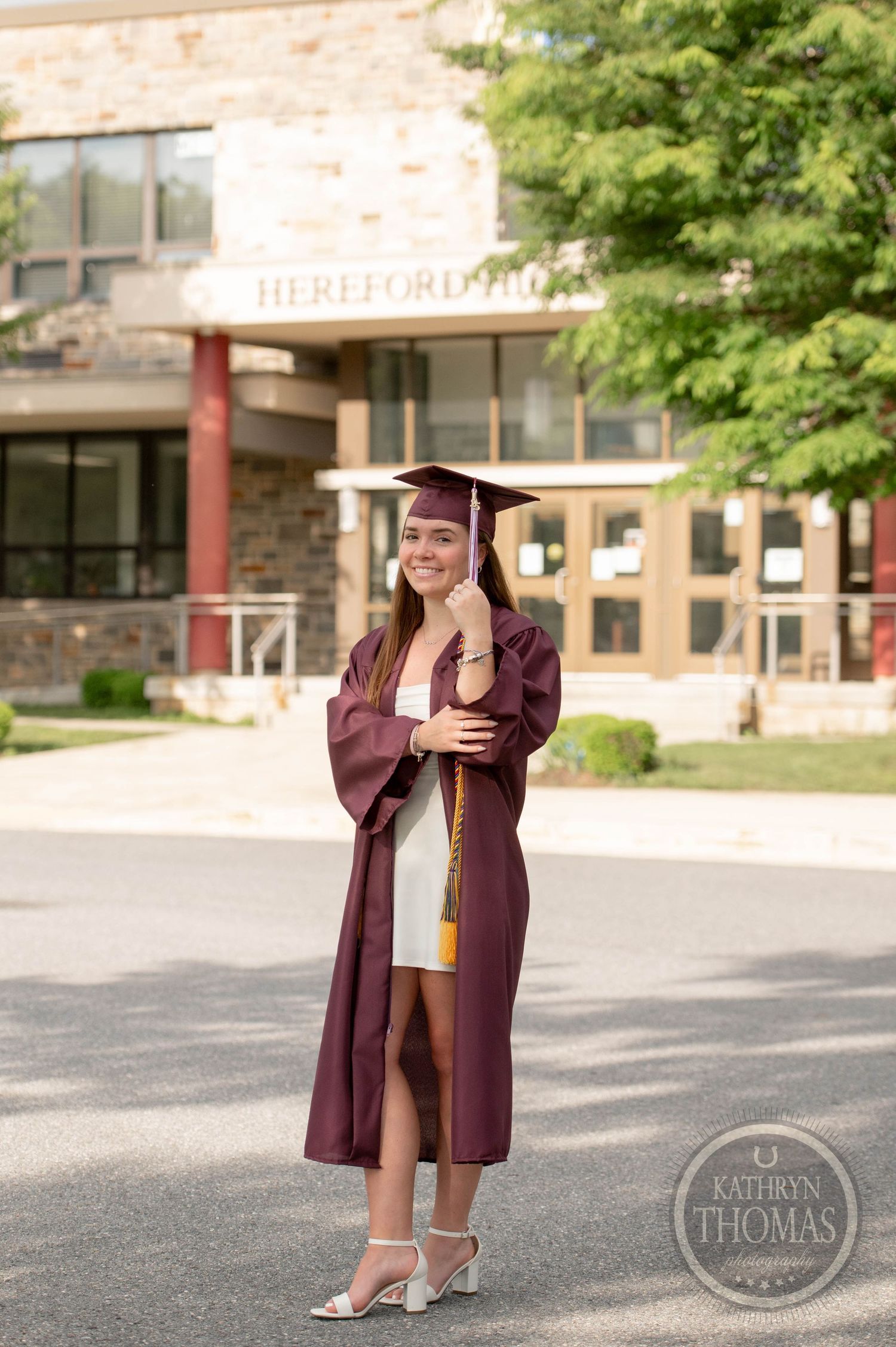 Graduate in maroon cap and gown poses outdoors in front of brick building during commencement ceremony.