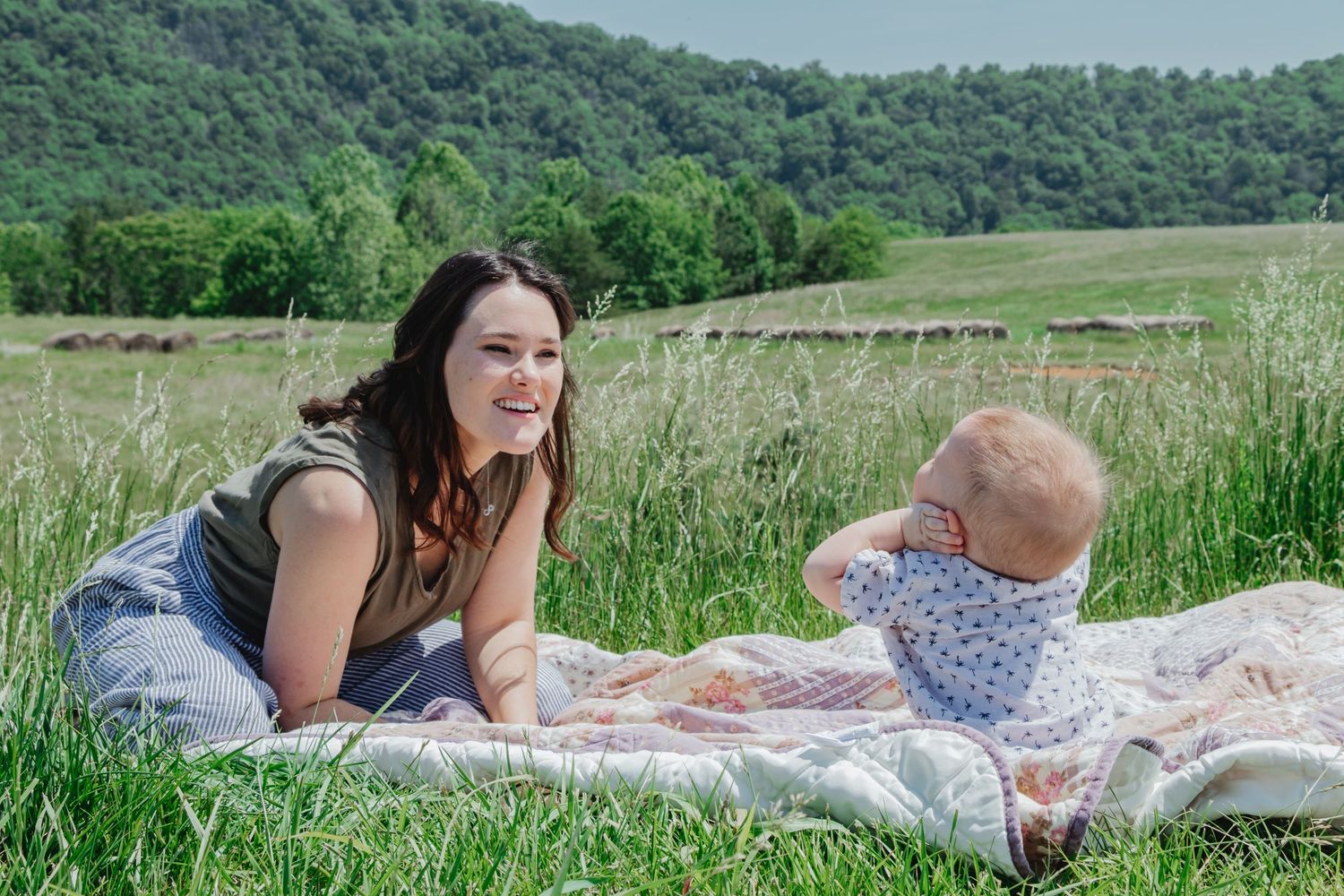 Mother smiling at her baby who is sitting on a quilt in the grass, looking up at the sky on a sunny day.