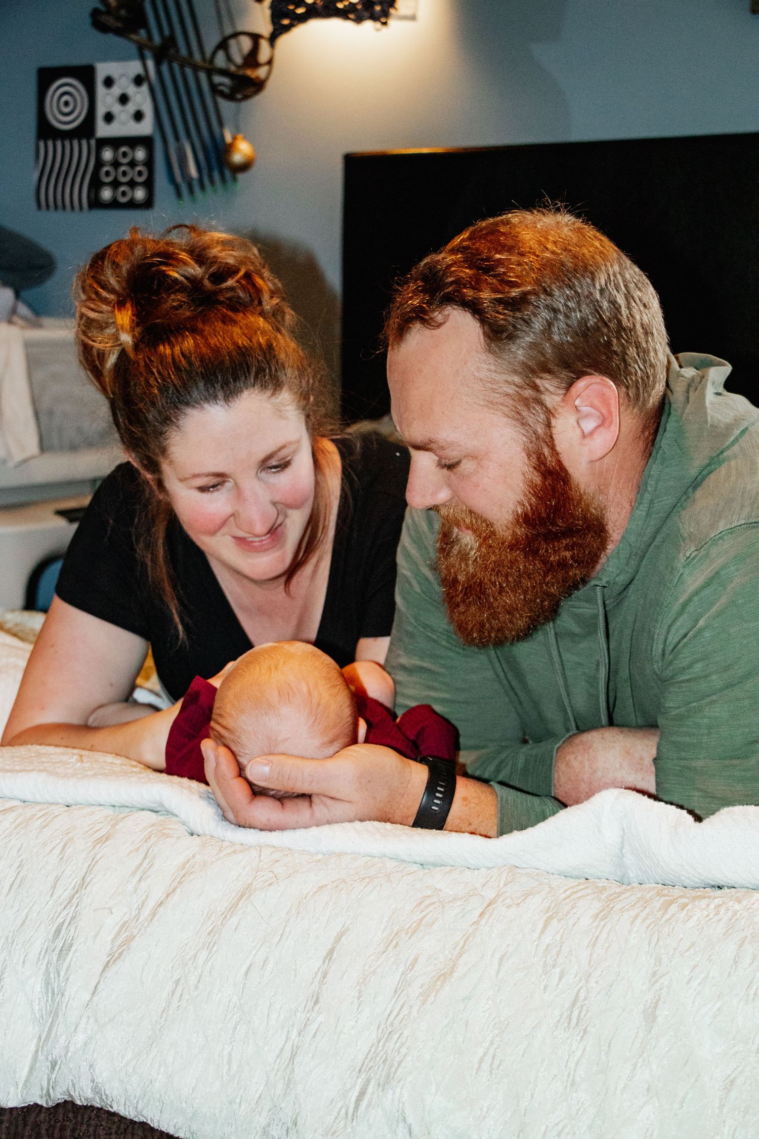 Mother & father lying on a bed lovingly gazing at their newborn, cradled gently in the father's hands.