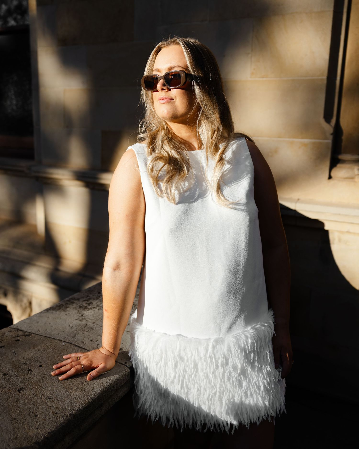 Stylish white sleeveless dress with feather trim detail captures golden sunlight while person leans against wall wearing sunglasses.