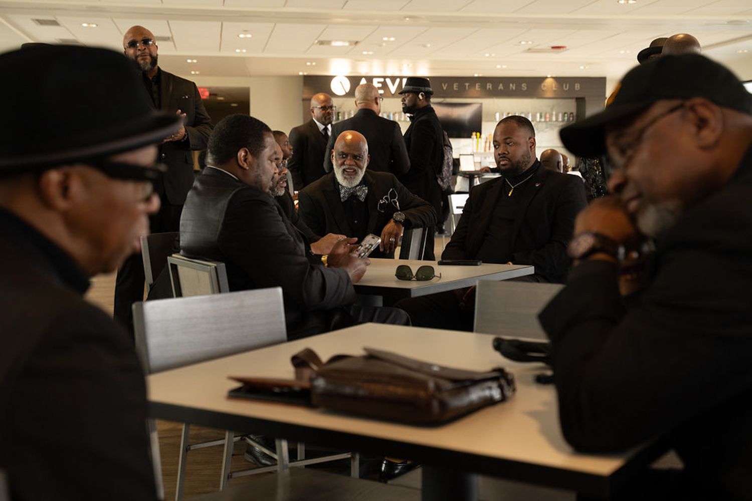 A group of black men seated around tables in a room wearing dark clothing and hats while having a discussion.