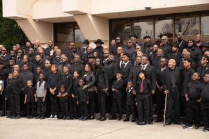 Large group of men and boys in formal black attire pose together on steps outside a building for a formal photograph.