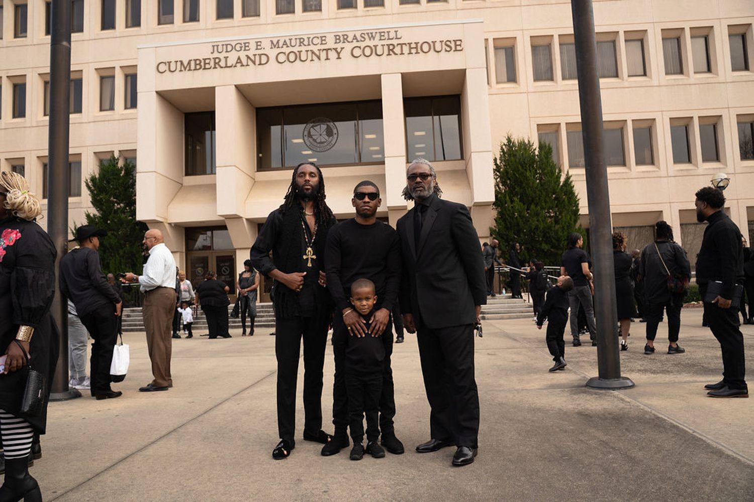 Three people stand together outside a courthouse building as others gather on the steps.