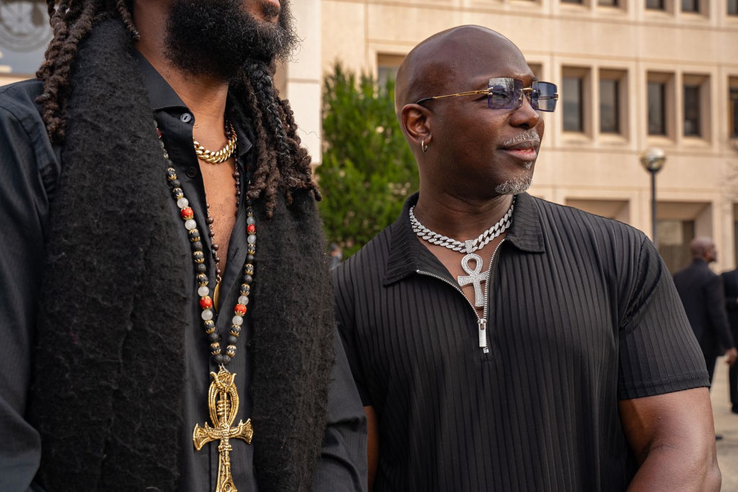 Two black men in contrasting outfits stand outside a courthouse with one wearing ankh and dark clothing.