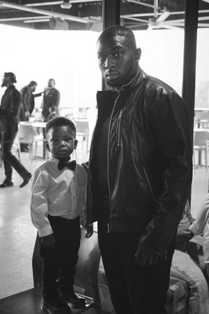 A black and white photograph showing an black man and young child standing together in a casual indoor setting.