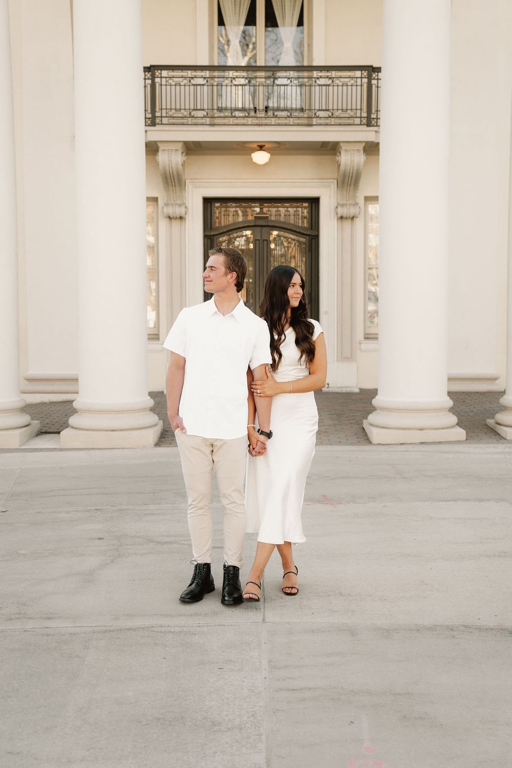 Elegant couple in white outfits standing together in front of pillared entrance.