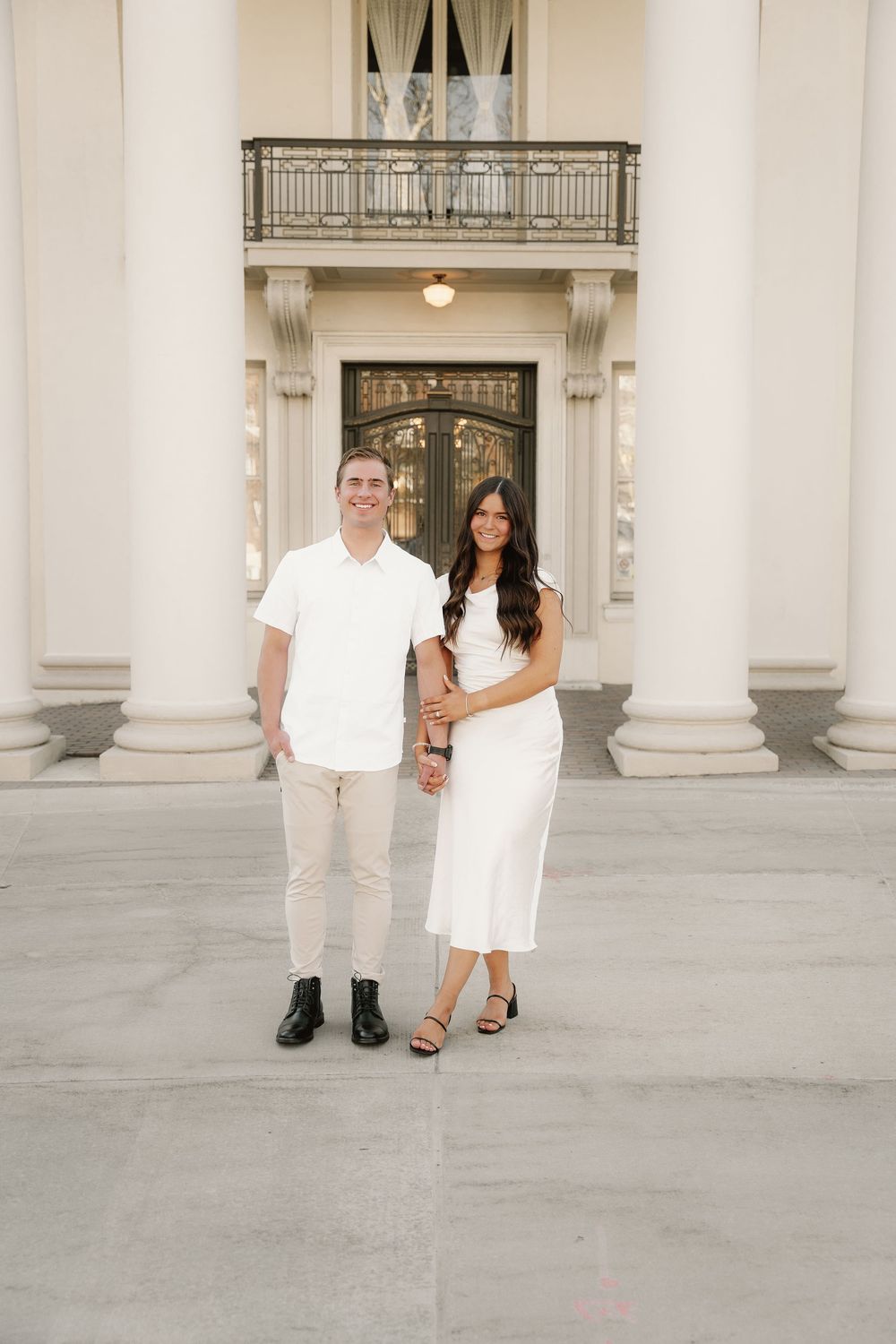 A couple in all-white attire stands together on a grand porch with white columns and an ornate doorway.