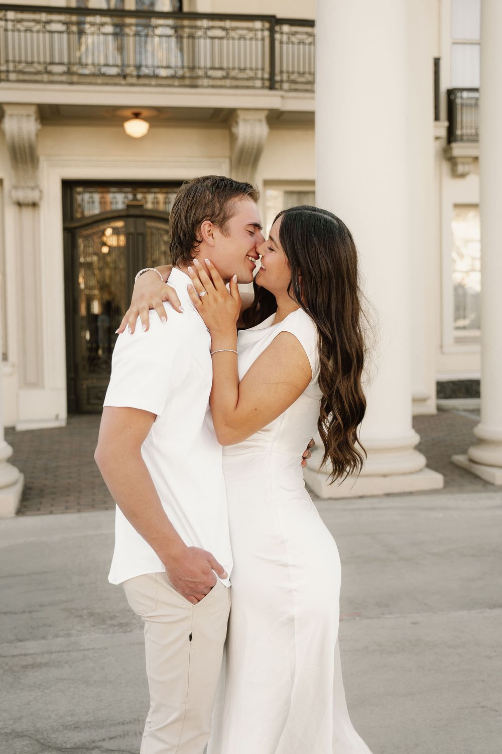 A couple dressed in white embraces and kisses during a romantic outdoor photoshoot at an elegant venue.
