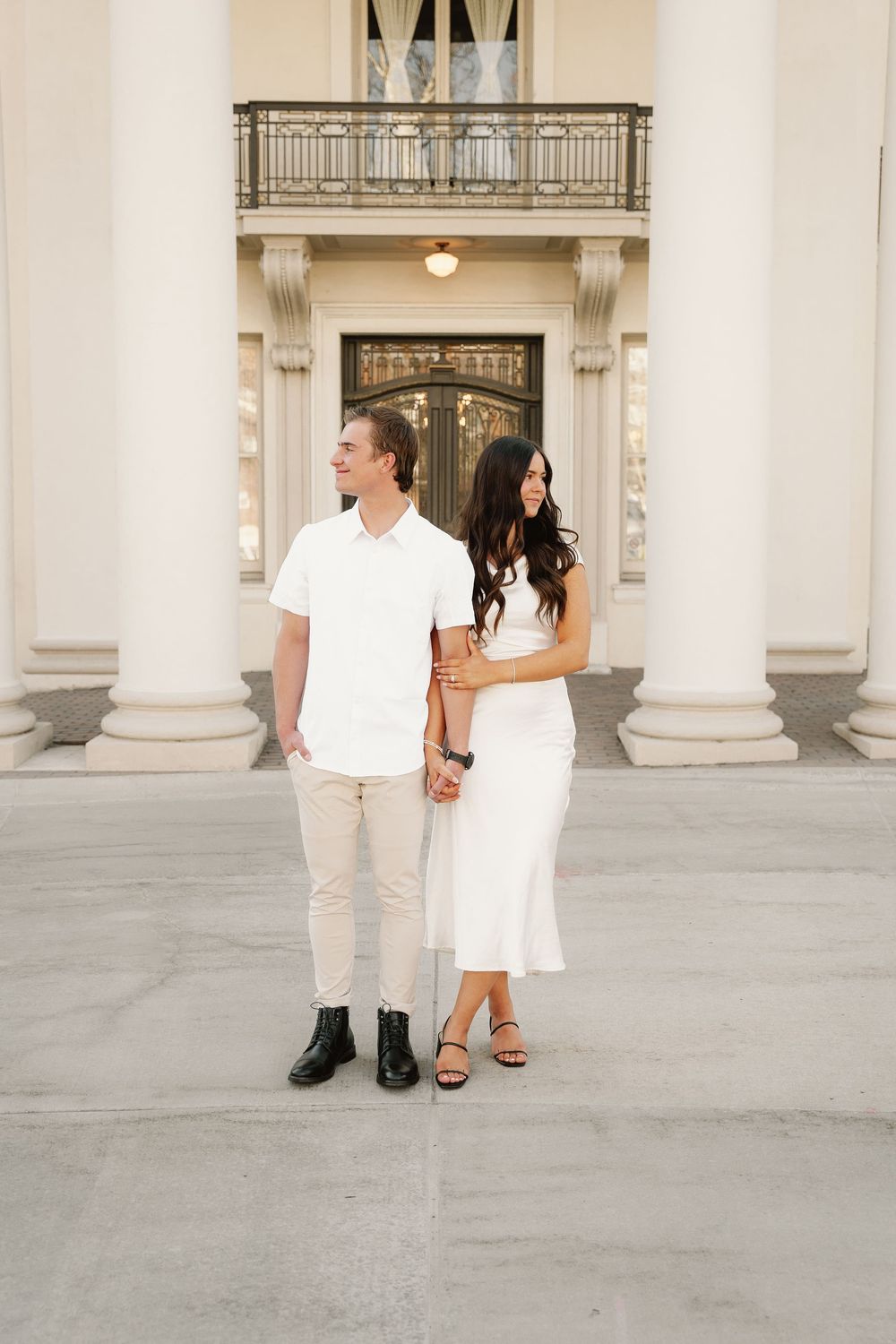 A couple in matching white attire holds hands while standing in front of a stately building entrance.