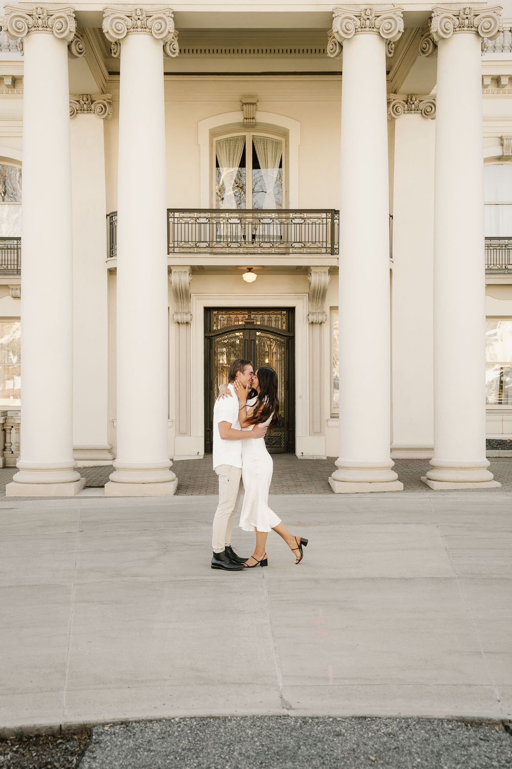 A couple in all white embraces while standing between grand Corinthian columns of a historic building.