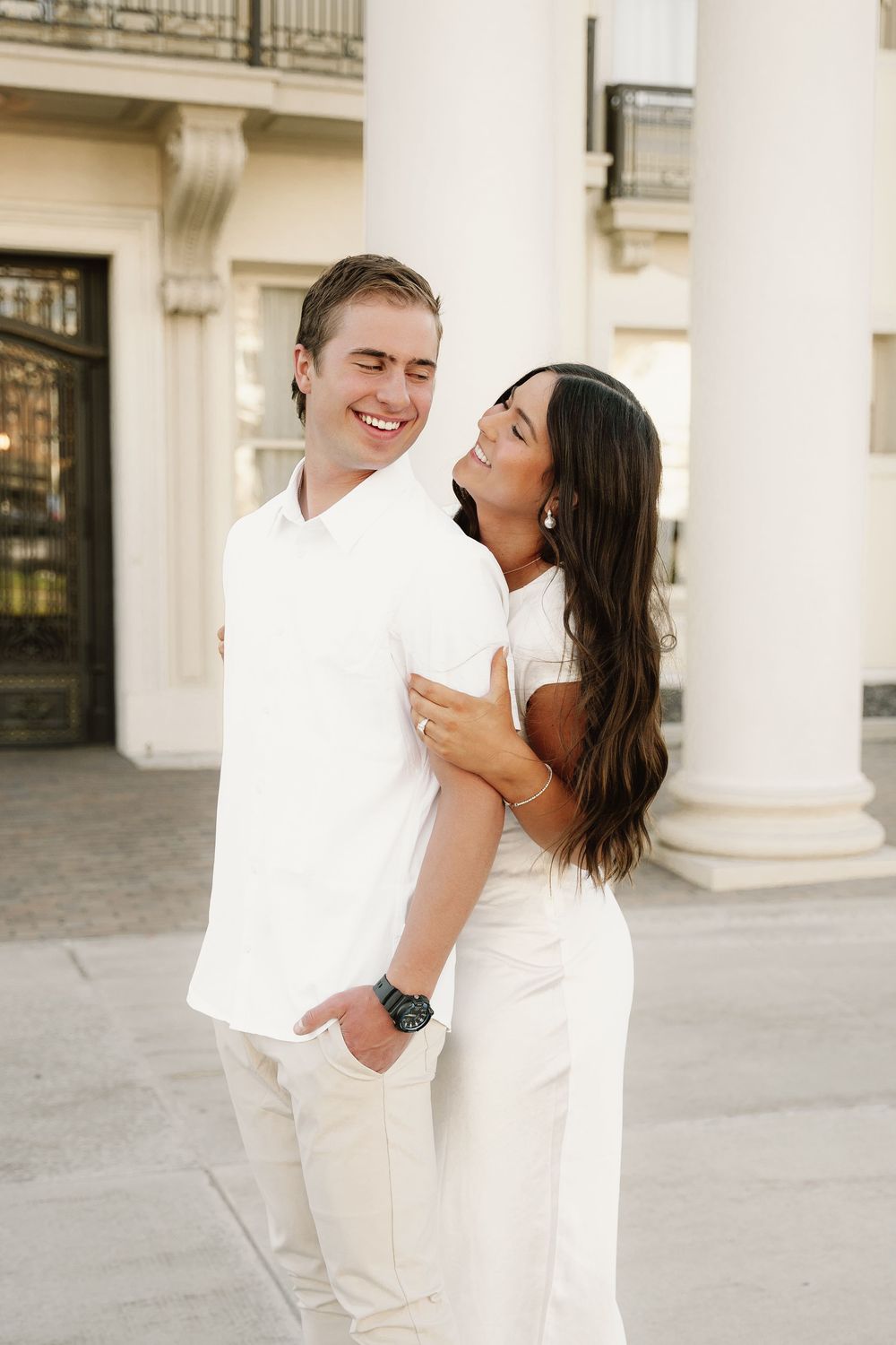Happy couple in white outfits laughing together against elegant architectural backdrop with columns.