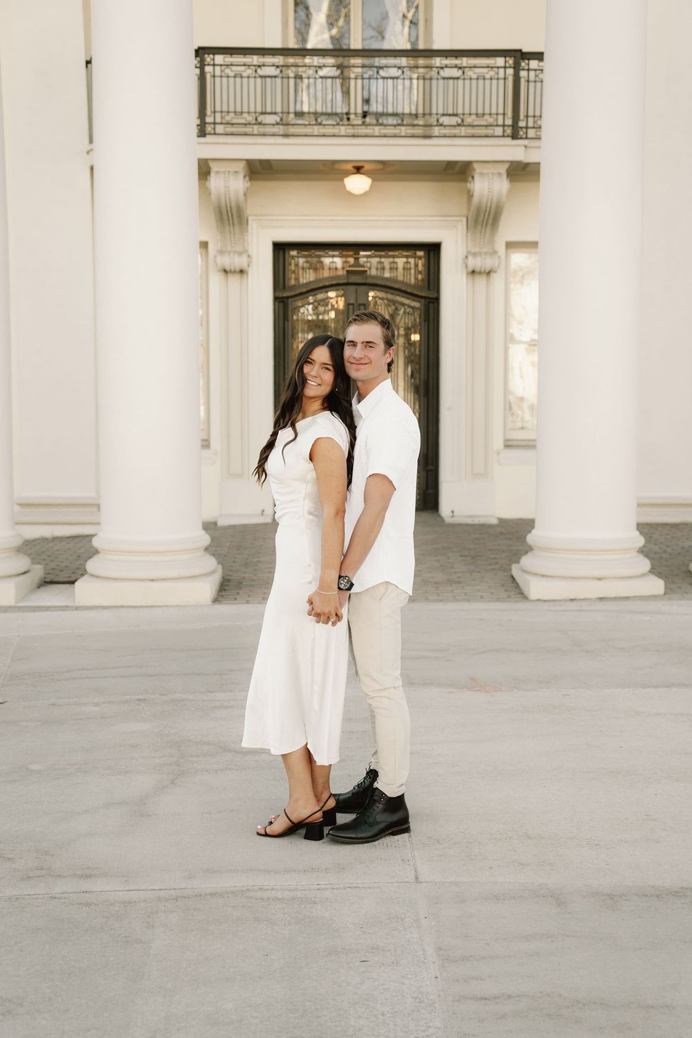 A couple in coordinated white outfits poses together in front of an elegant entrance with white columns.