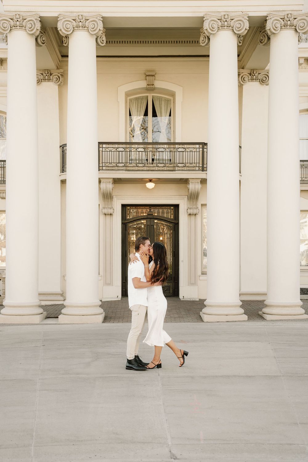 A couple shares a romantic moment in white outfits against a backdrop of grand classical architecture.