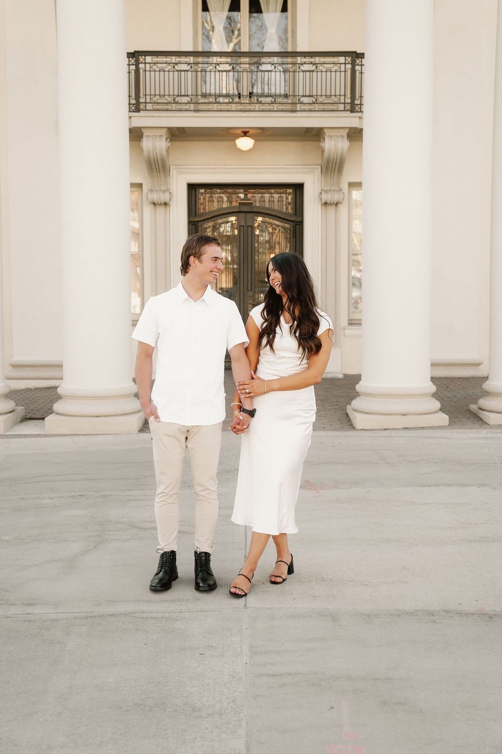 Two people in white outfits stand together in front of ornate columns and a decorative door at an elegant venue.