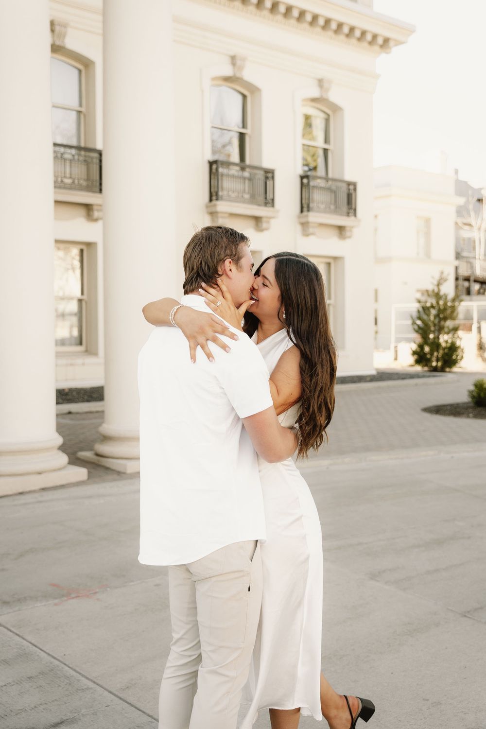 A romantic couple in white outfits share an intimate moment outside a classical building with ornate balconies.
