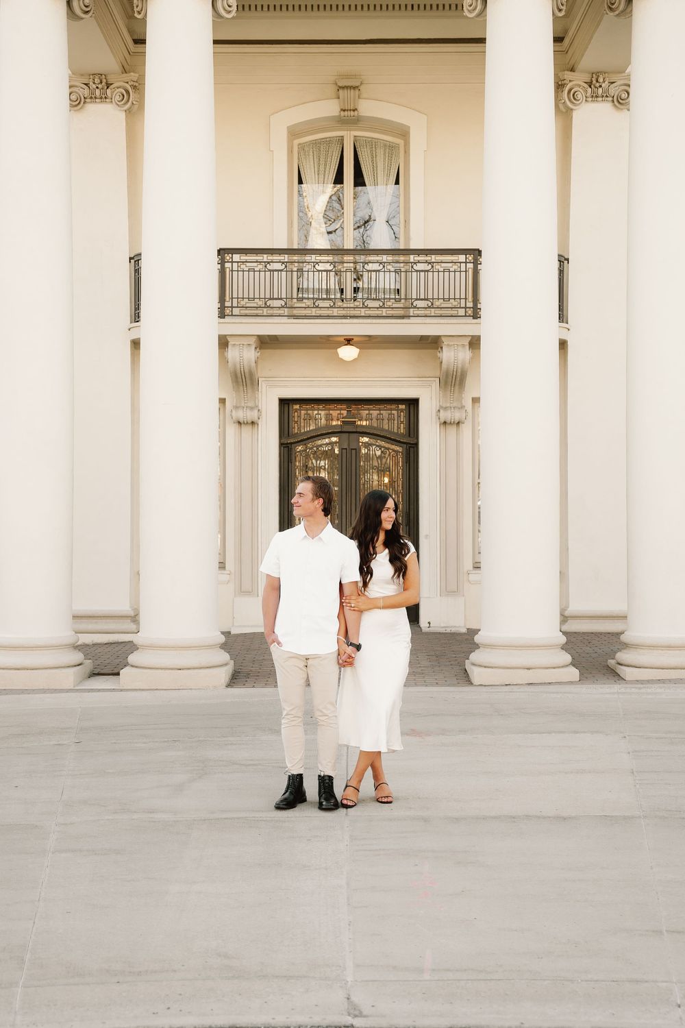 A couple in all-white outfits stands in front of an elegant building with classical white columns and ornate architecture.