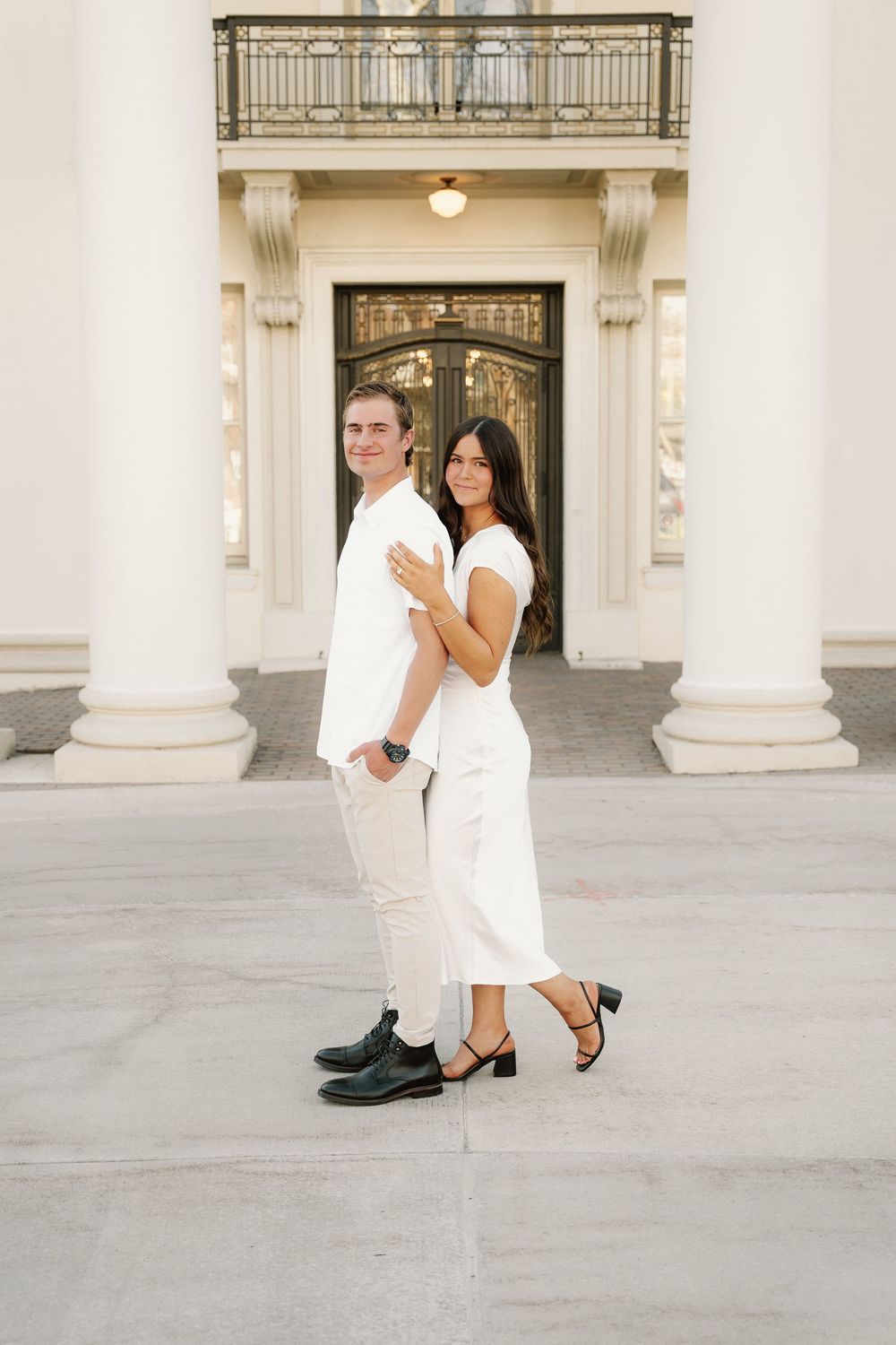 Couple in coordinated white outfits pose back-to-back against pillared building entrance.