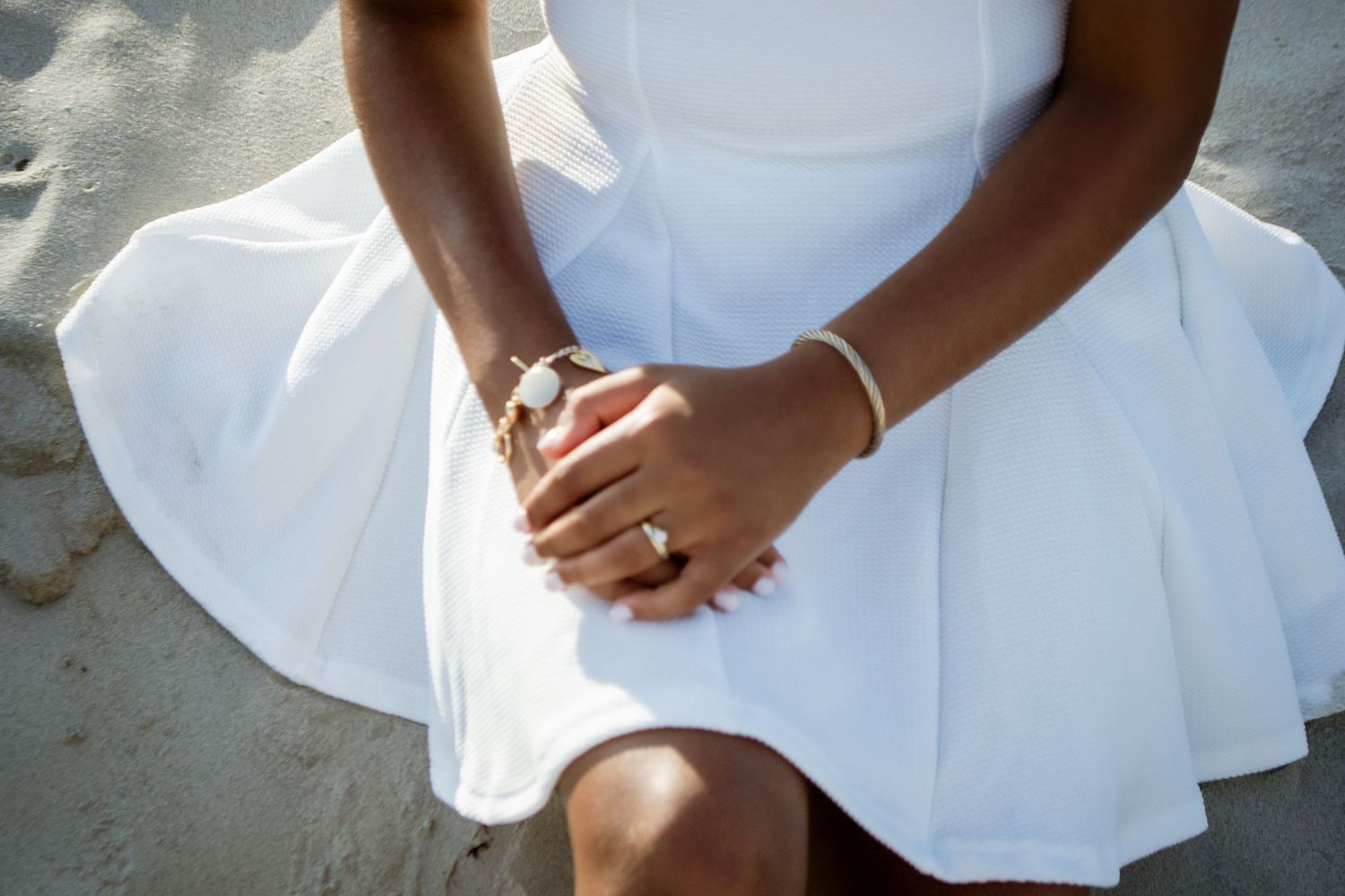 Close-up of hands folded in lap wearing white dress in sunlight.