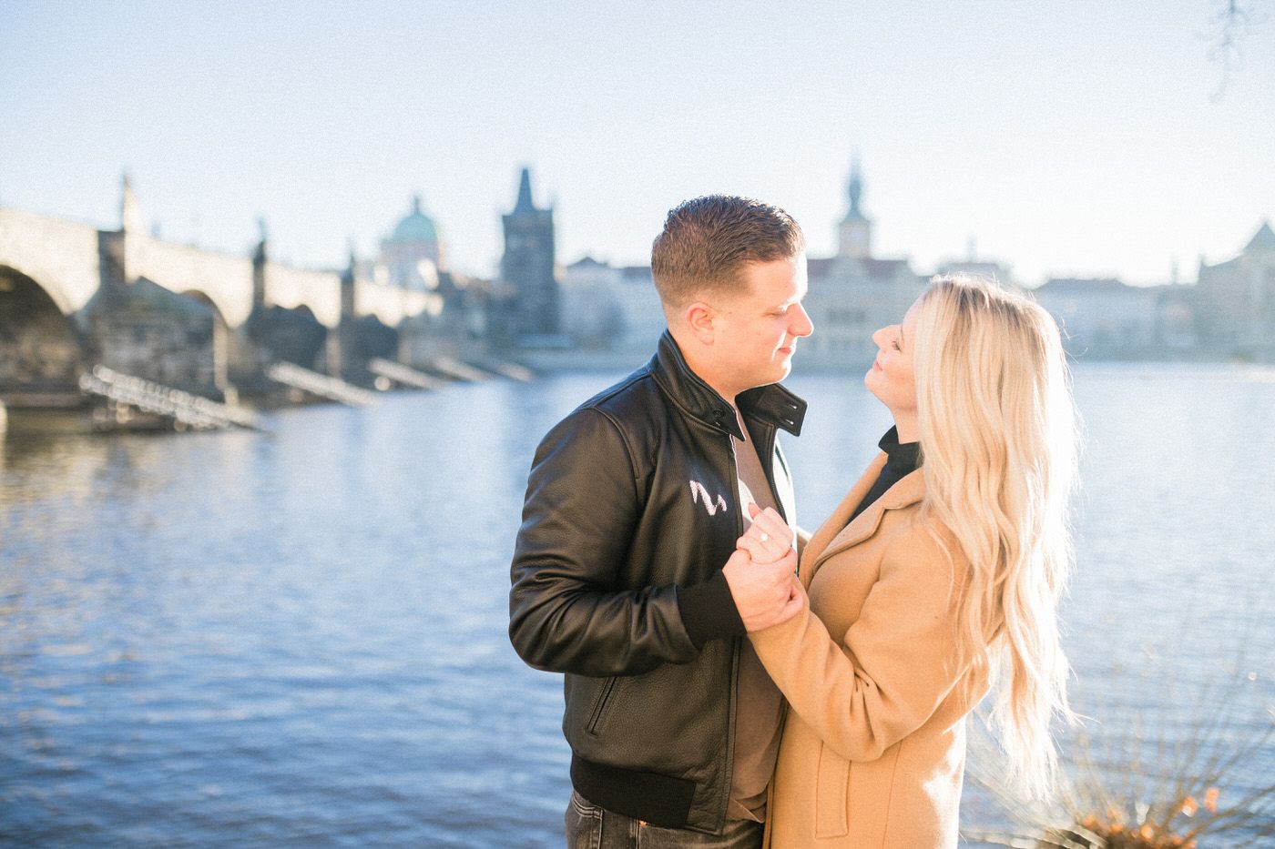 A couple in winter coats stands romantically by a river with a historic European cityscape Prague and Charles bridge in the background.