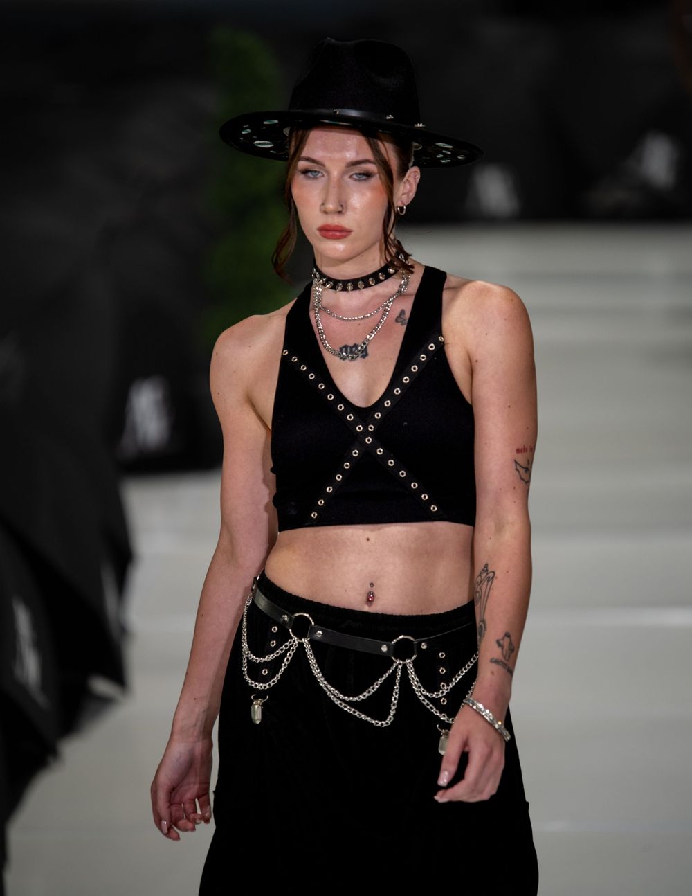 Alternative fashion model wearing black crop top with chains and wide brim hat on catwalk.