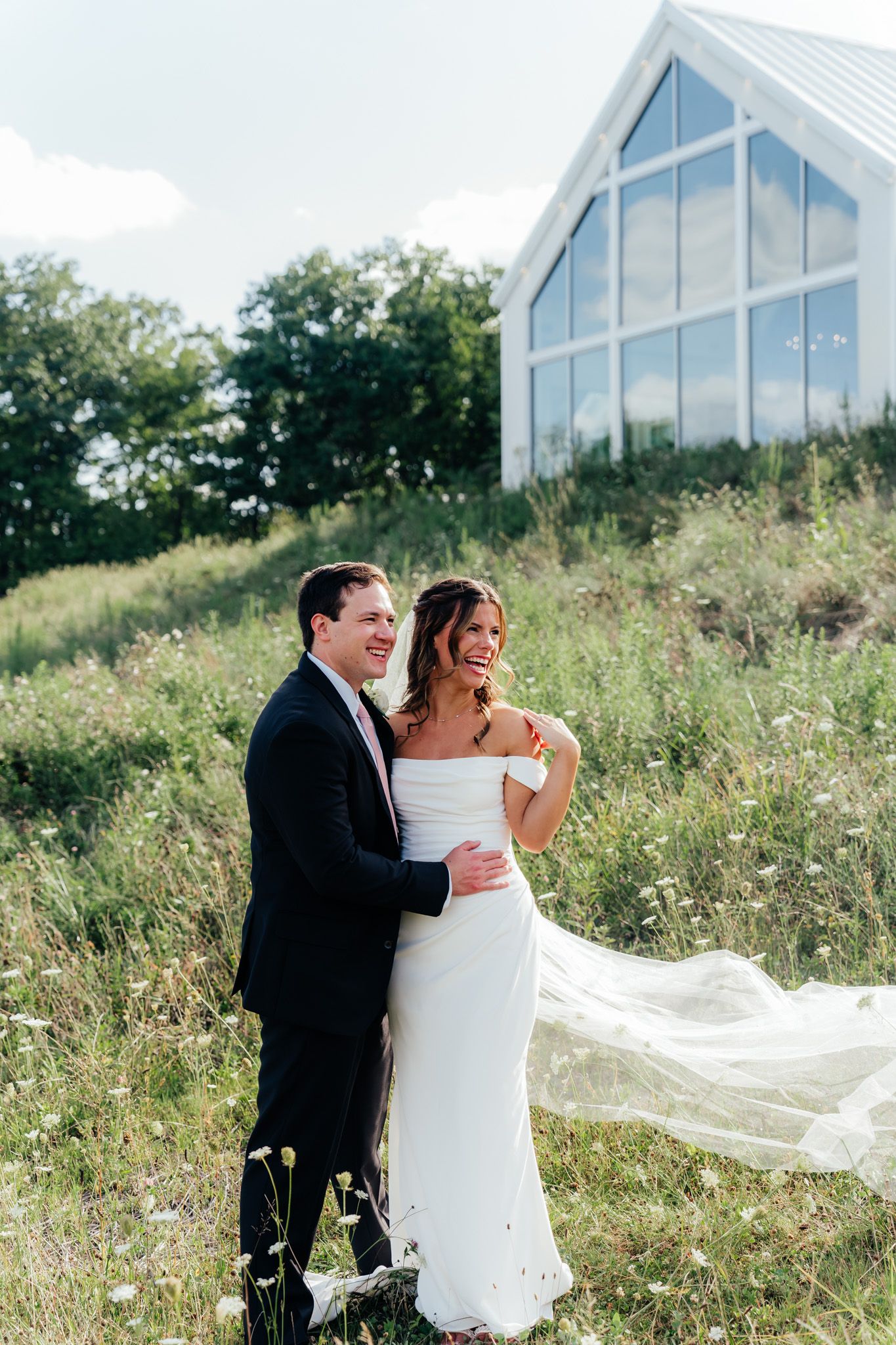 A couple in wedding attire embraces on a grassy hillside with a modern white chapel in the background.