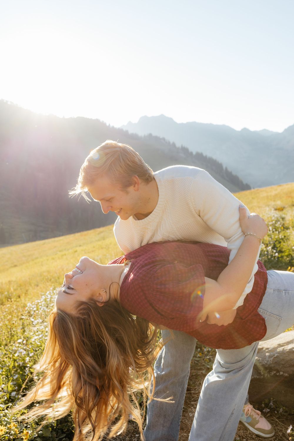 Man dipping laughing woman in a sunny alpine meadow with mountain backdrop.