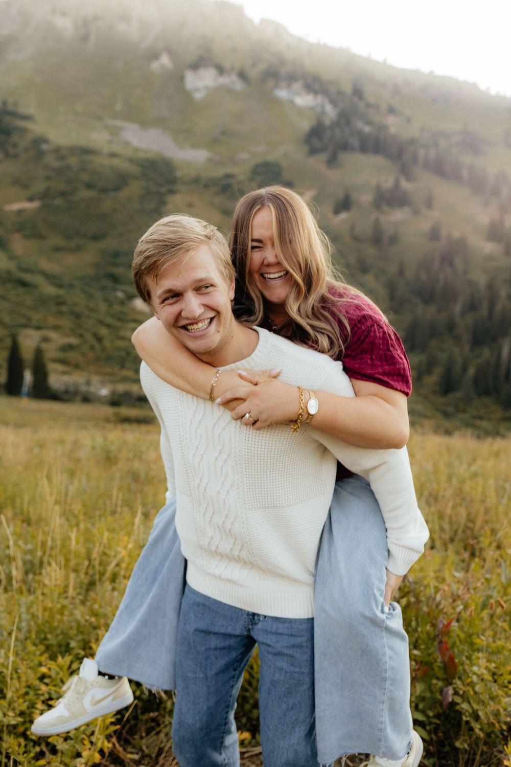 Woman piggyback riding laughing man in a lush mountain meadow landscape.