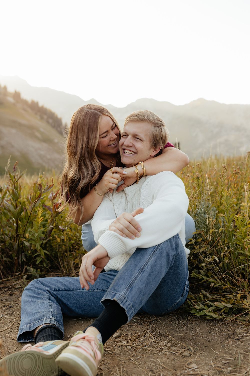 A woman hugs a smiling man from behind while sitting in a mountain wildflower field.