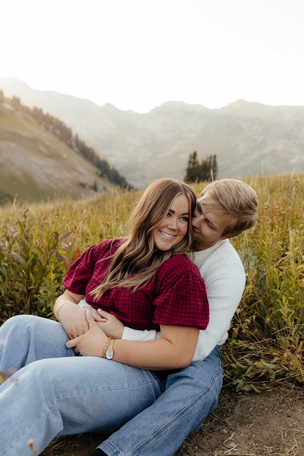 A couple snuggles together in a sunlit mountain meadow, woman in red top smiling.