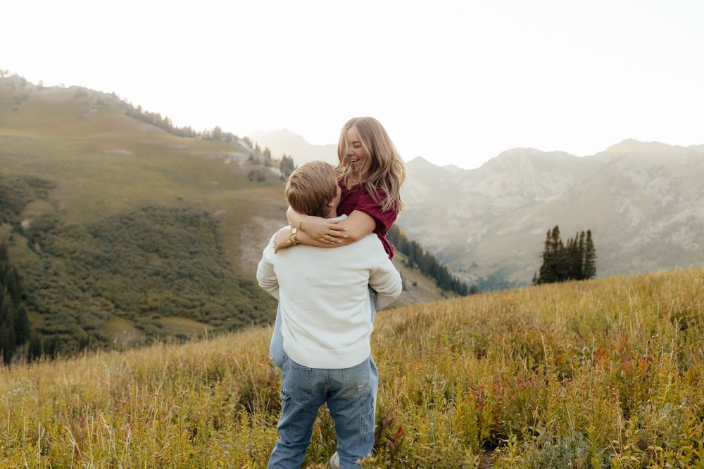 A man lifts a laughing woman in a red top in a scenic mountain meadow.
